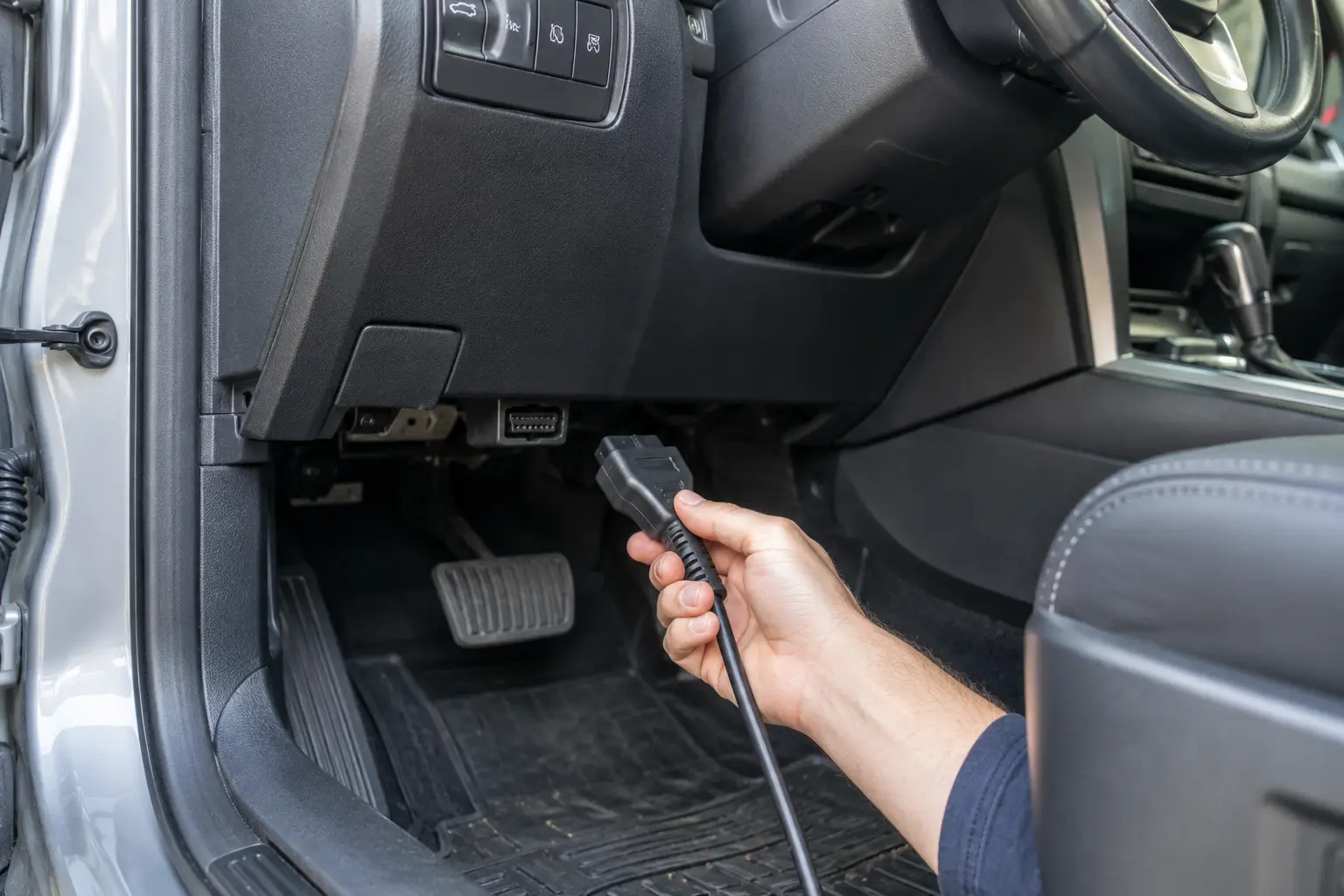 A hand holding an OBD2 connector near the diagnostic port under a car dashboard.