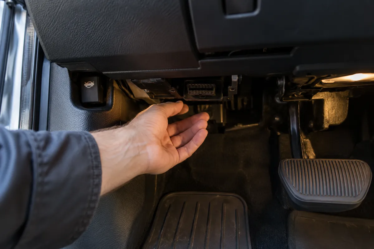 Mechanic reaching beneath the driver's side dashboard to locate the OBD2 diagnostic port.