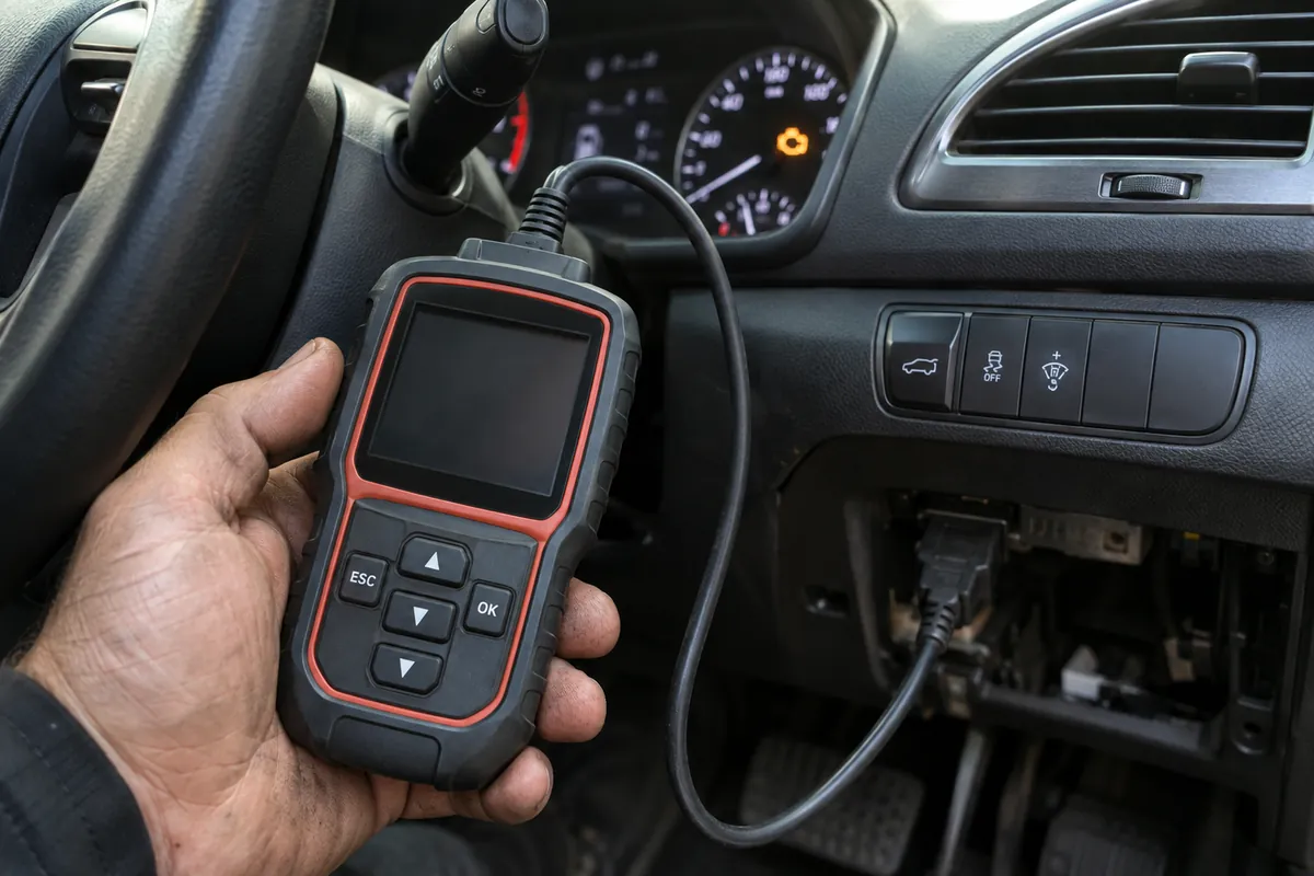Mechanic holding an OBD2 scanner with a blank screen connected to a car's diagnostic port while the check engine light glows on the dashboard.