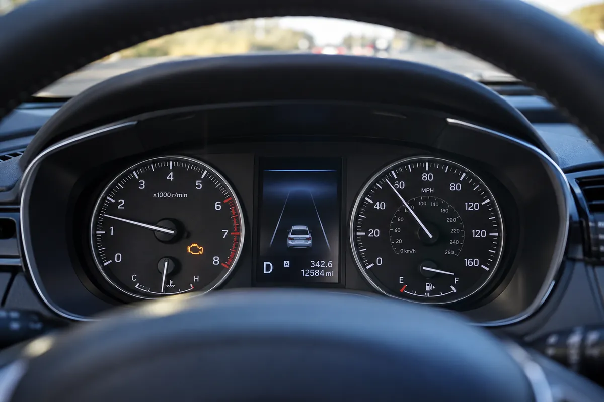Modern car dashboard showing an illuminated amber check engine light, analog speedometer with MPH markings, and tachometer while the engine is running.